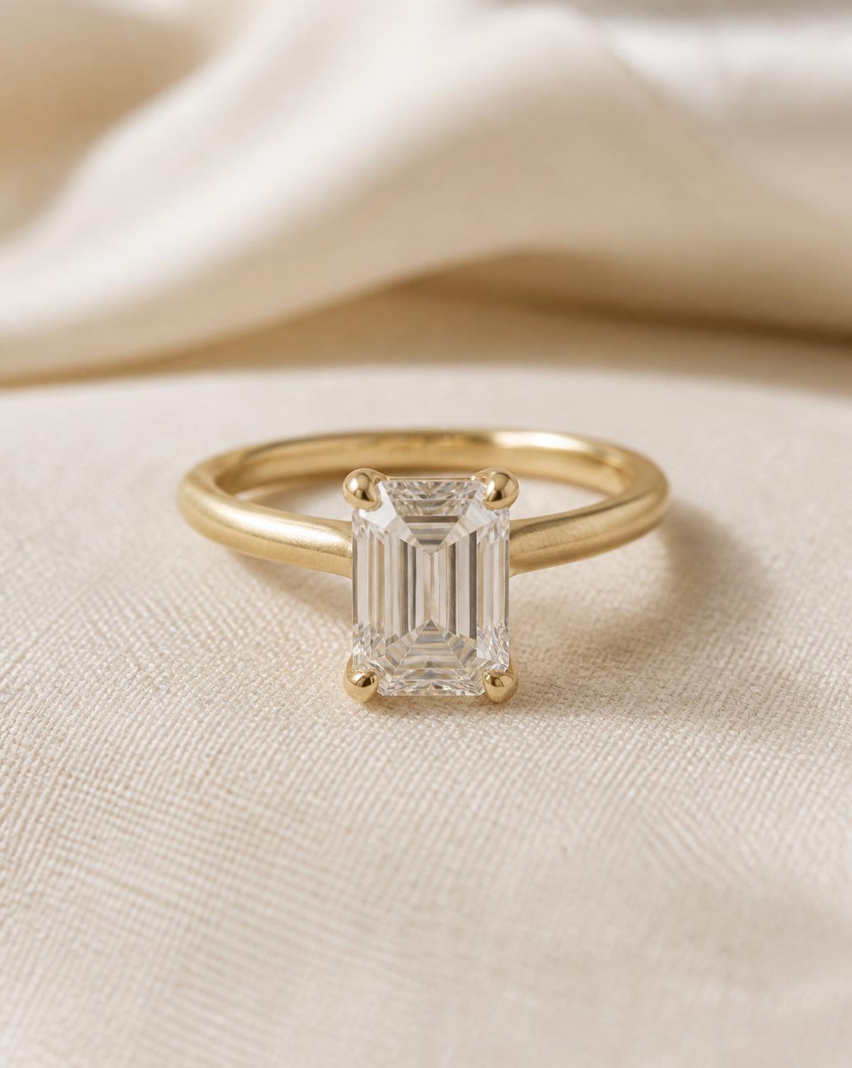 Gold solitaire ring with an emerald cut diamond resting on champagne linen